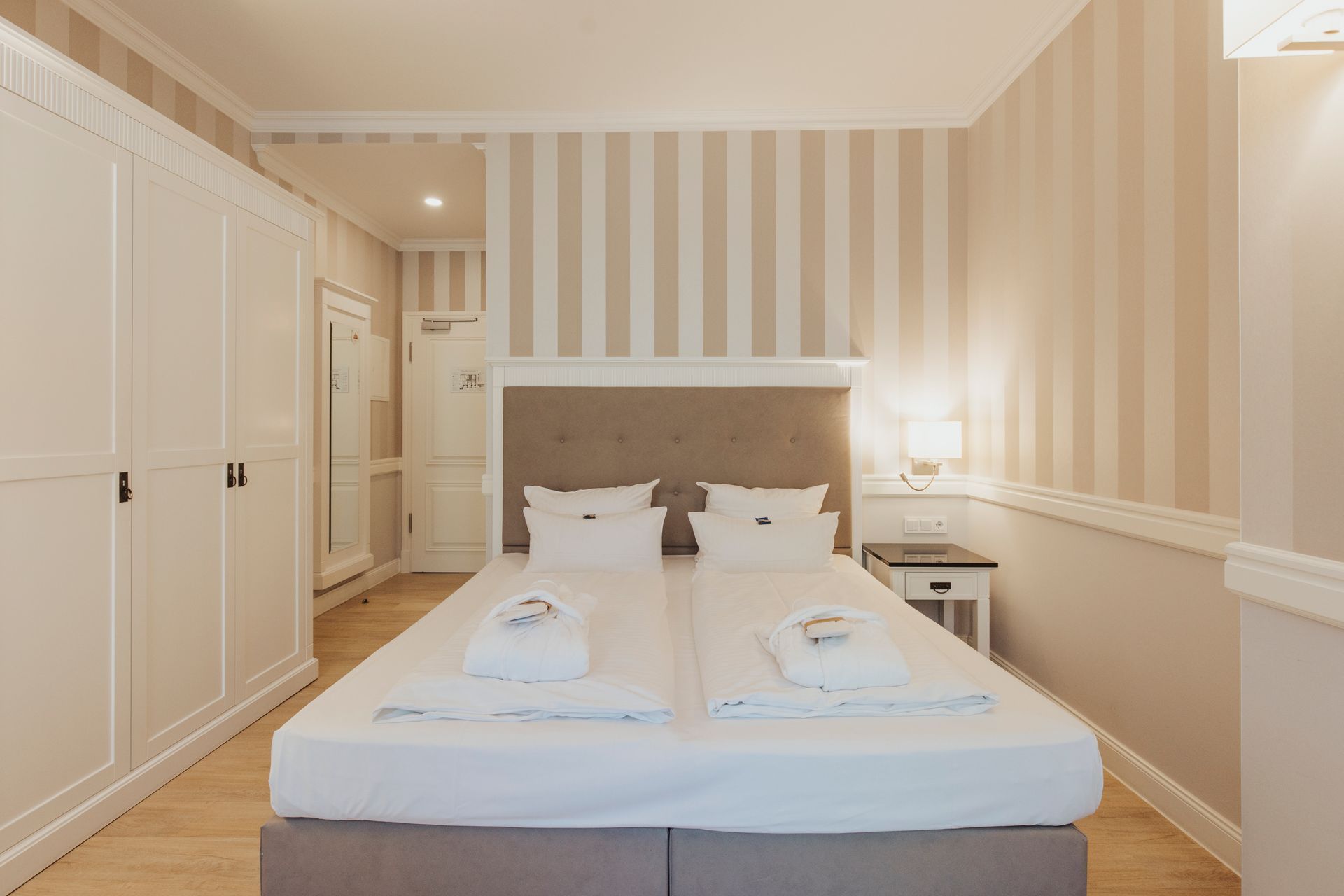 Helles Schlafzimmer mit ordentlich gemachtem Bett, gestreifter Tapete, weißem Kleiderschrank und Nachttischlampe