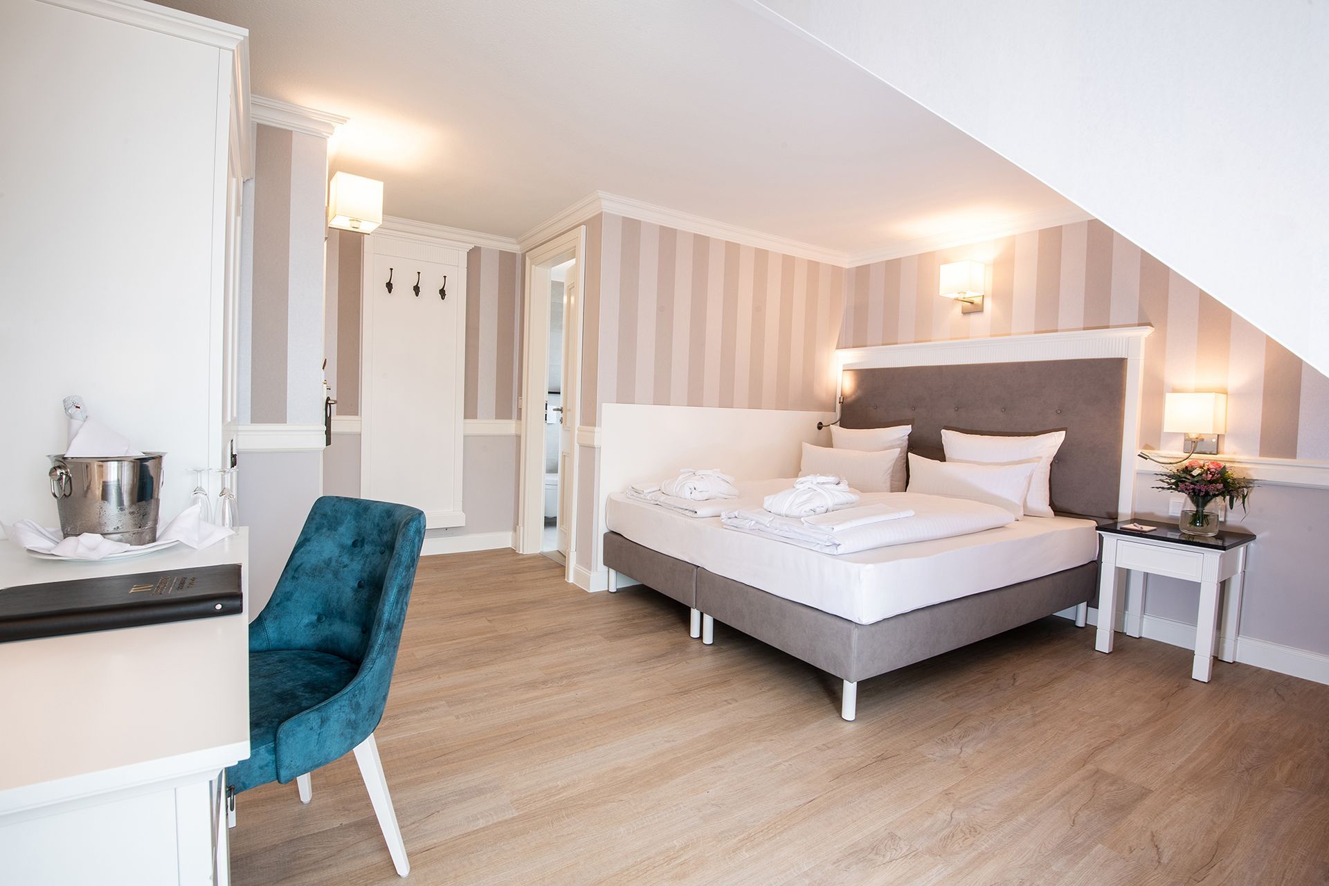 Helles, modernes Schlafzimmer mit gestreifter Tapete, türkisfarbenem Sessel und einem ordentlich gemachten Bett mit weißer Bettwäsche.