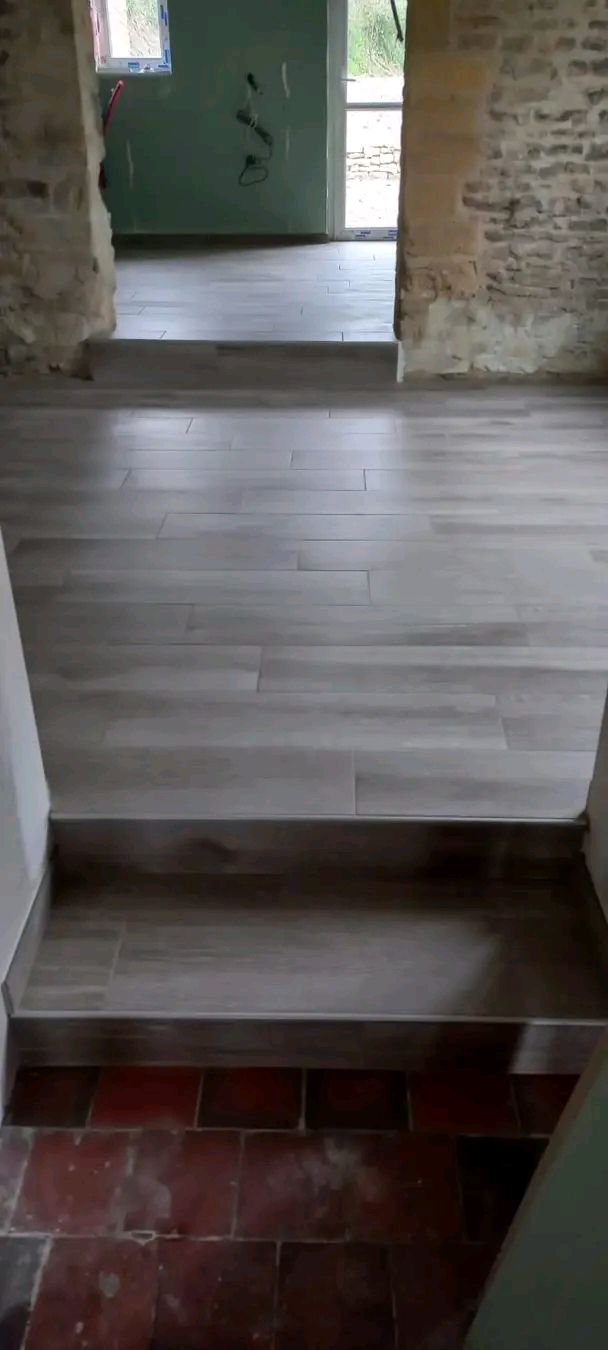 Une marche mène à un parquet clair, jouxtant des carreaux rouges, donnant sur une porte dans une pièce inachevée.