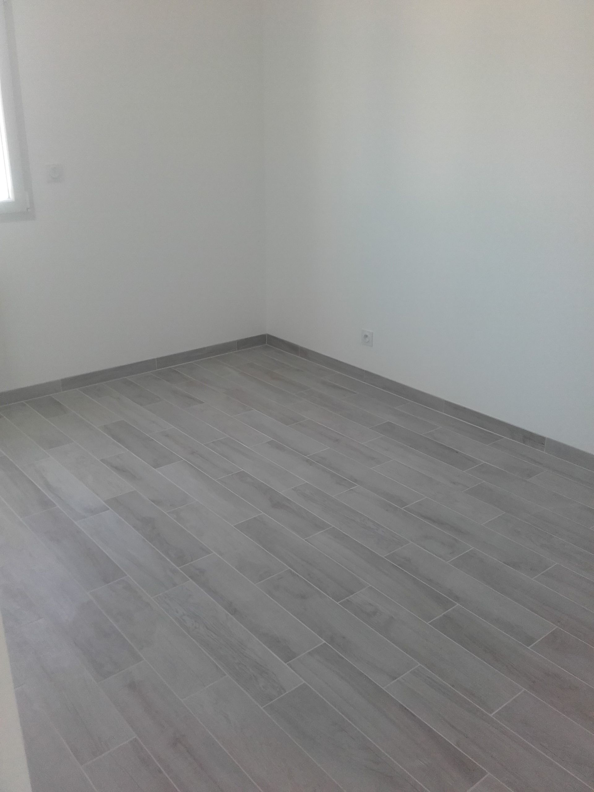 Pièce vide avec parquet gris, murs blancs et une fenêtre sur la gauche.