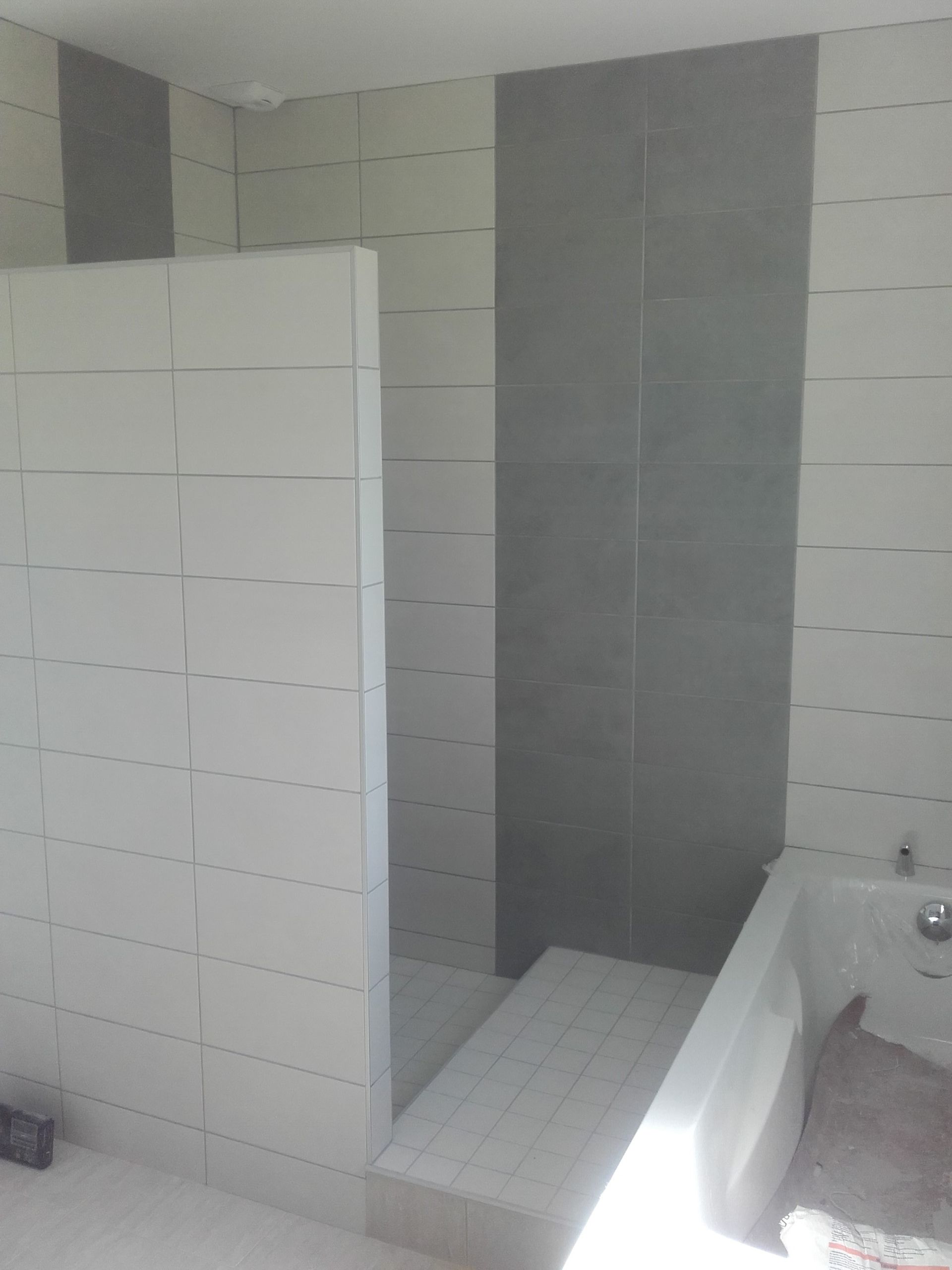 Salle de bains partiellement carrelée avec murs blancs et gris, un coin douche et un rebord de baignoire sur la droite.