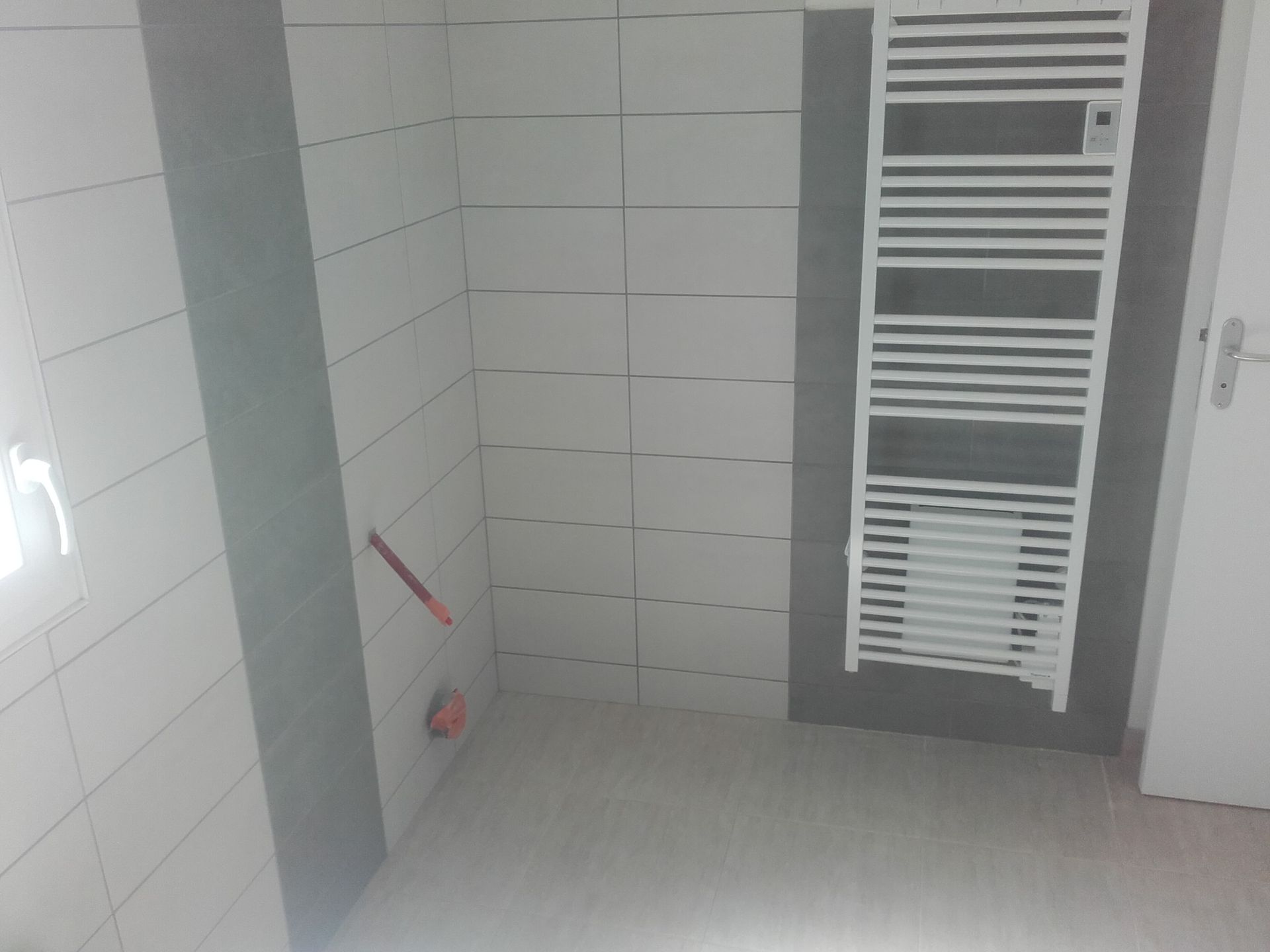 Petite salle de bains carrelée avec un sèche-serviettes blanc, une porte et des produits de nettoyage dans un coin.