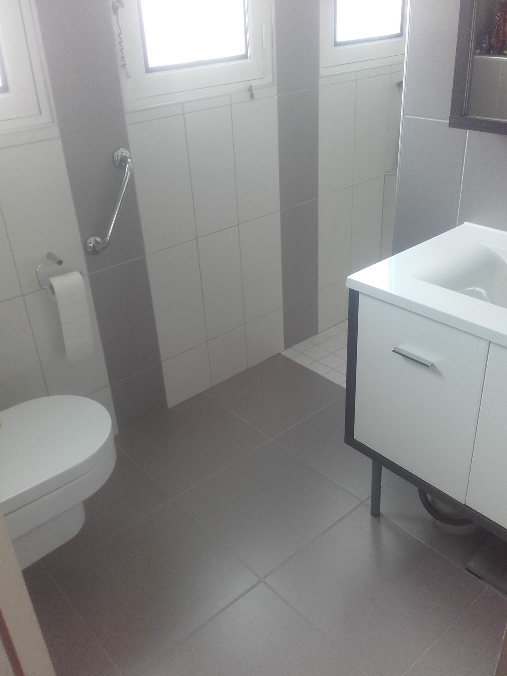 Petite salle de bains avec toilettes, fenêtre, murs carrelés, lavabo et sol gris.