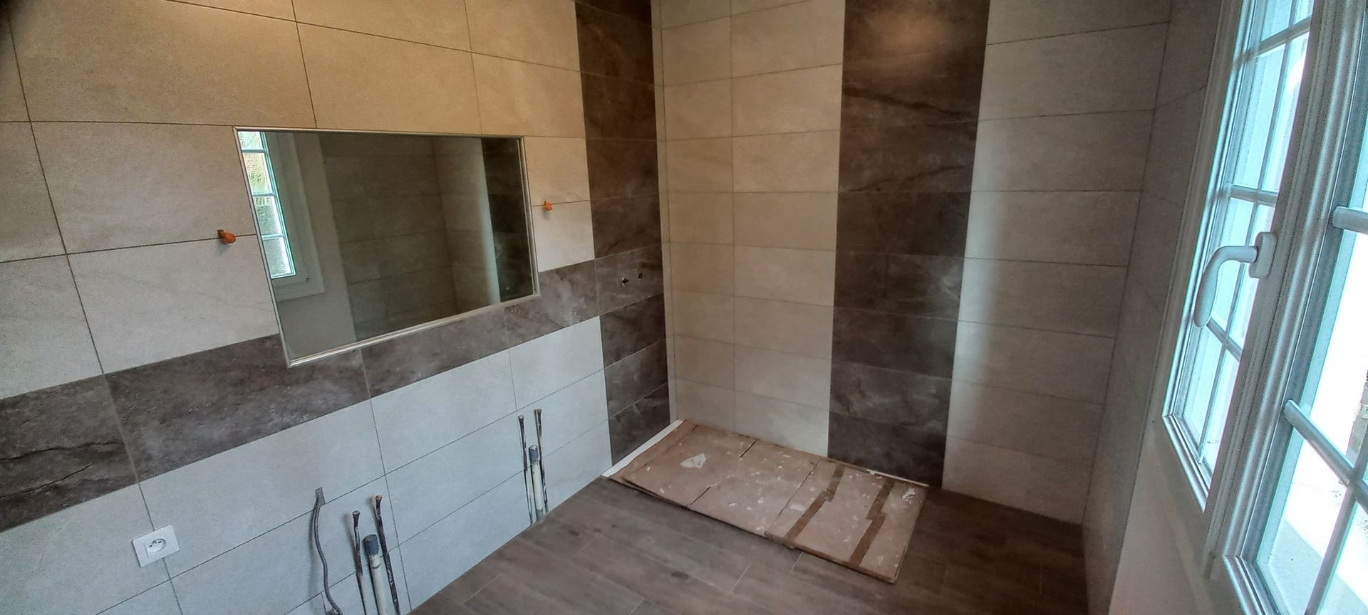 Salle de bain partiellement carrelée avec un grand miroir, un espace lavabo et de la lumière naturelle provenant d'une fenêtre près de la douche.