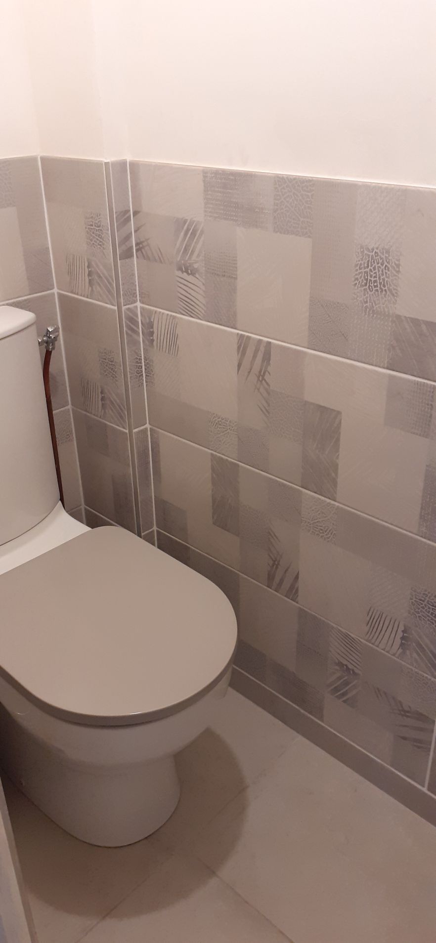 Petite salle de bains avec des toilettes blanches à côté d'un mur et d'un sol carrelés gris clair.