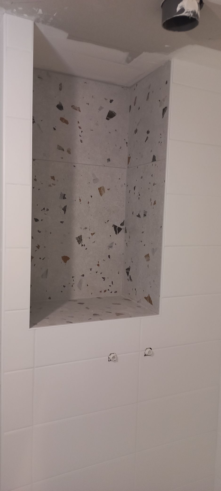Mur de douche carrelé de blanc avec une étagère encastrée contenant des points noirs et une pomme de douche au-dessus