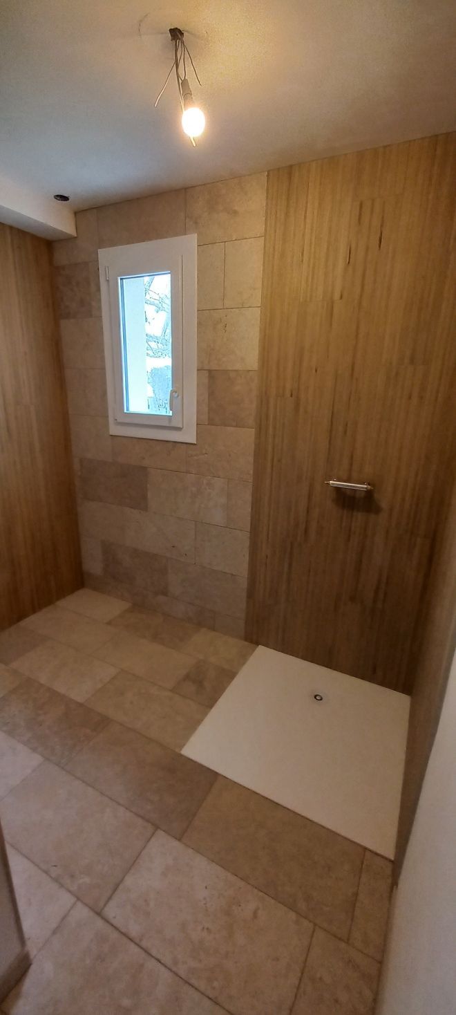 Petite salle de bains avec murs lambrissés, sol carrelé, fenêtre et espace douche.