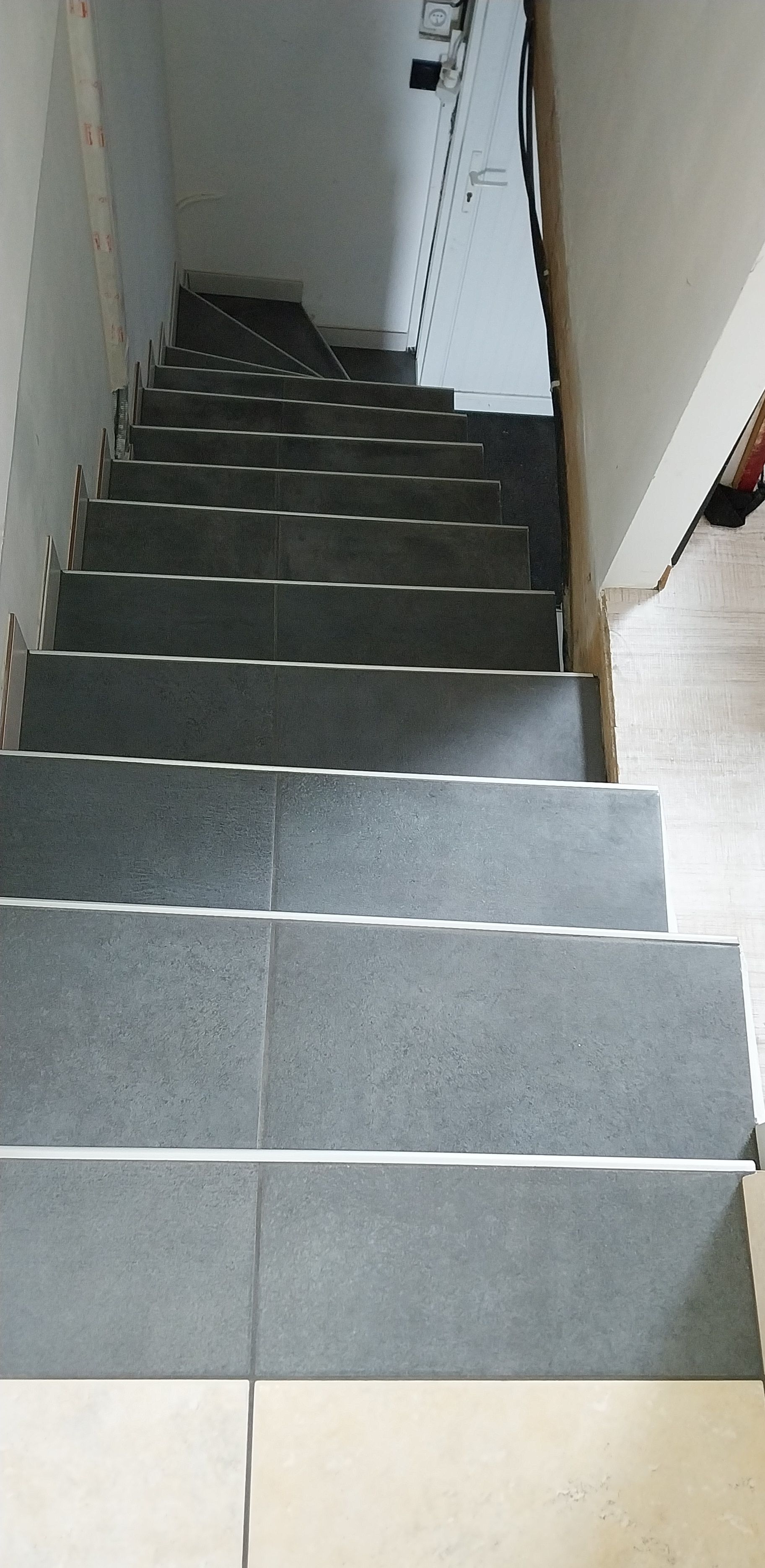 Escalier raide recouvert de moquette grise, avec des murs blancs et une rampe blanche, vu d'en bas.