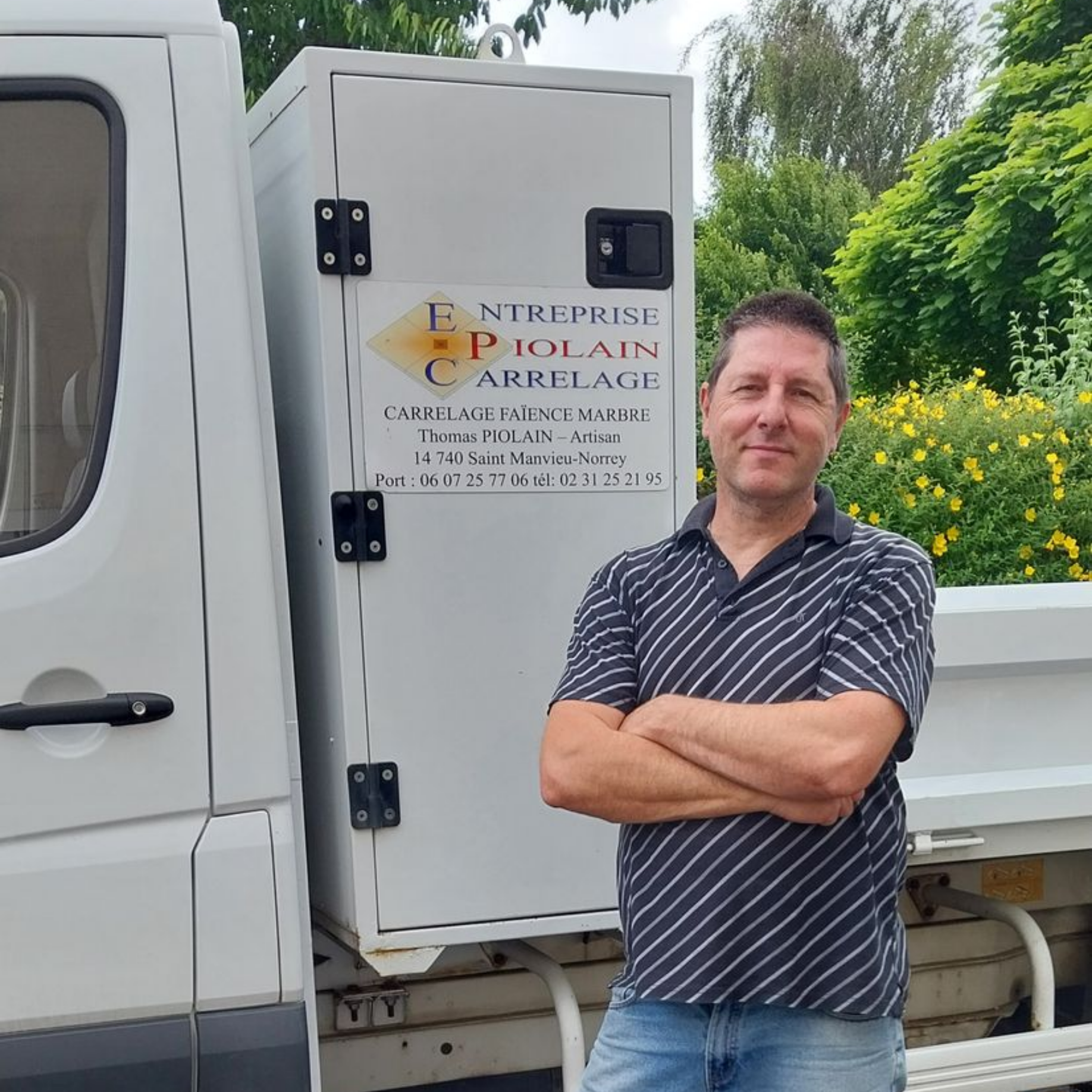 Un homme se tient debout, les bras croisés, à côté d'un camion blanc dans un cadre extérieur verdoyant.
