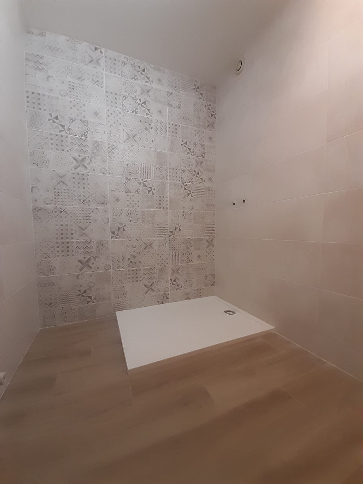 Salle de bain vide en cours de rénovation avec mur carrelé à motifs, receveur de douche blanc et sol imitation bois.