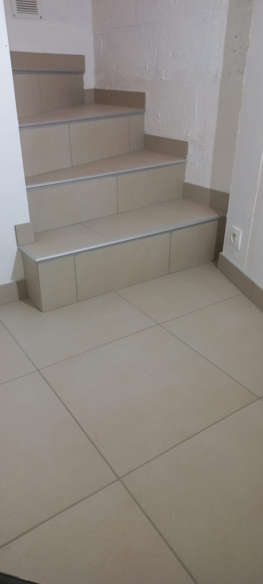 Escalier intérieur avec marches carrelées beiges et murs blancs, le long d'un sol carrelé.