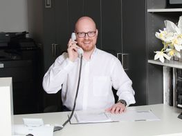 Hr. Neuhaus lächelnd beim Schreibtisch sitzend am Telefon