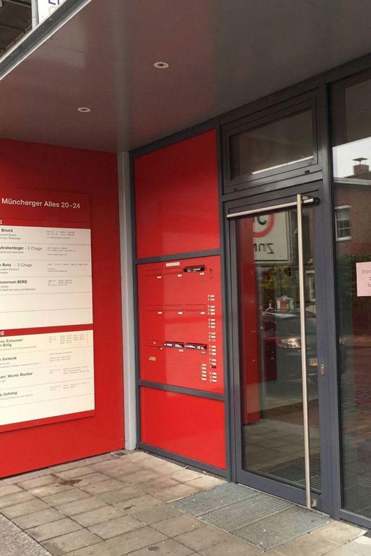 Roter Eingangsbereich mit Glastüren, roten Wänden und Aushängen unter einer überdachten Markise.