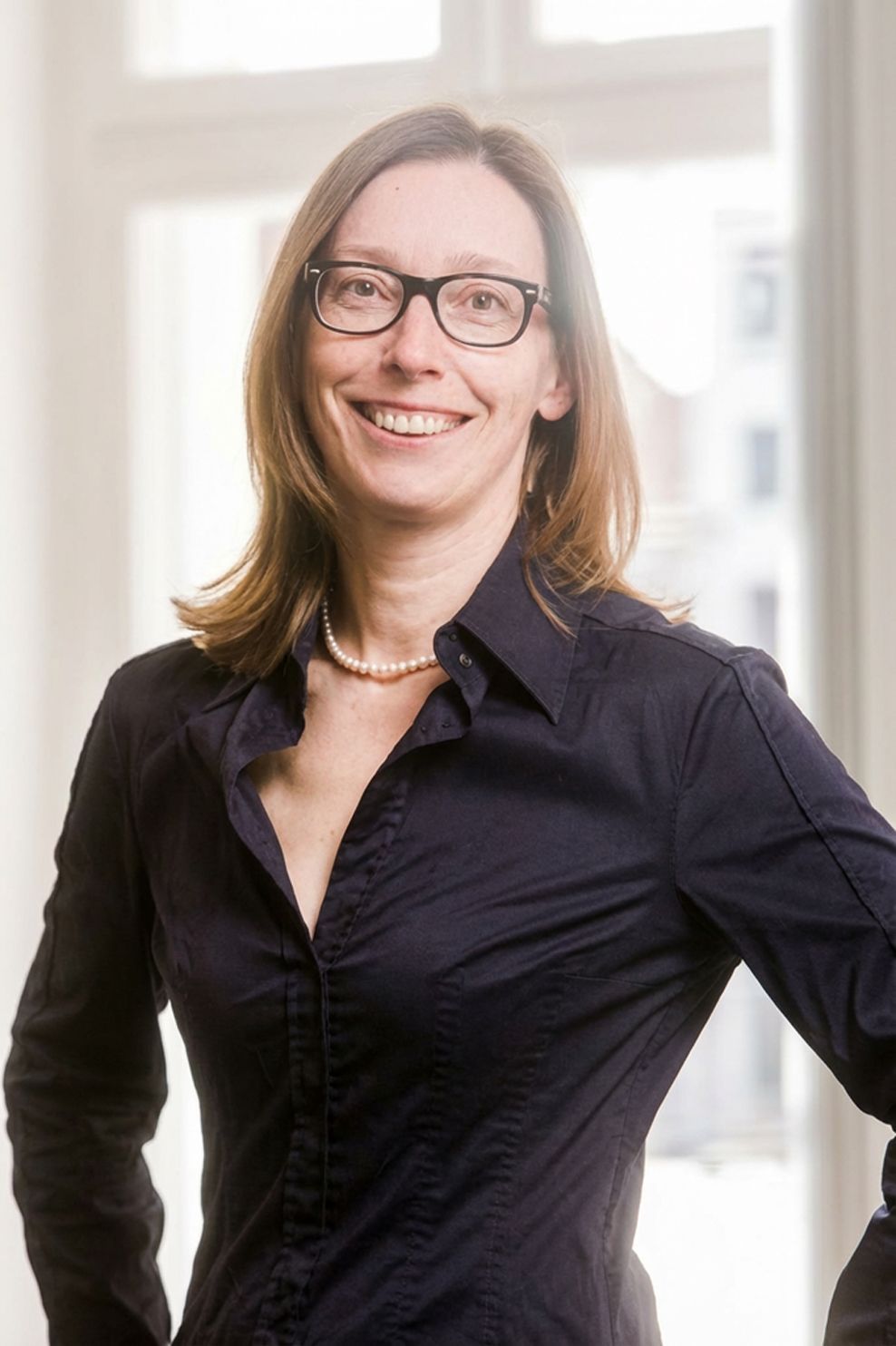Eine lächelnde Frau in dunkler Bluse und Brille steht an einem hellen Fenster.