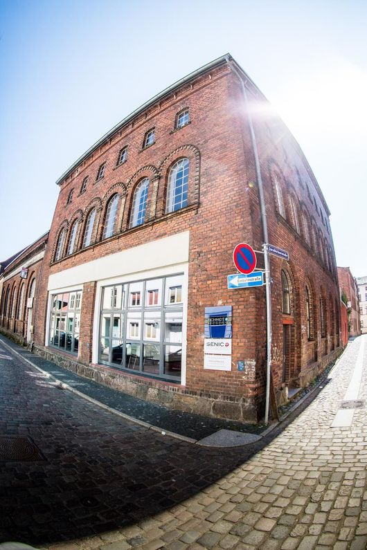 Eckgebäude aus rotem Backstein mit großen Fenstern an einer Kopfsteinpflasterstraße unter hellem Sonnenlicht