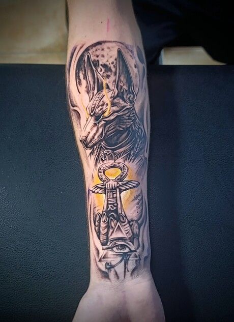 Una persona tiene un tatuaje de un lobo y un ankh en su antebrazo.