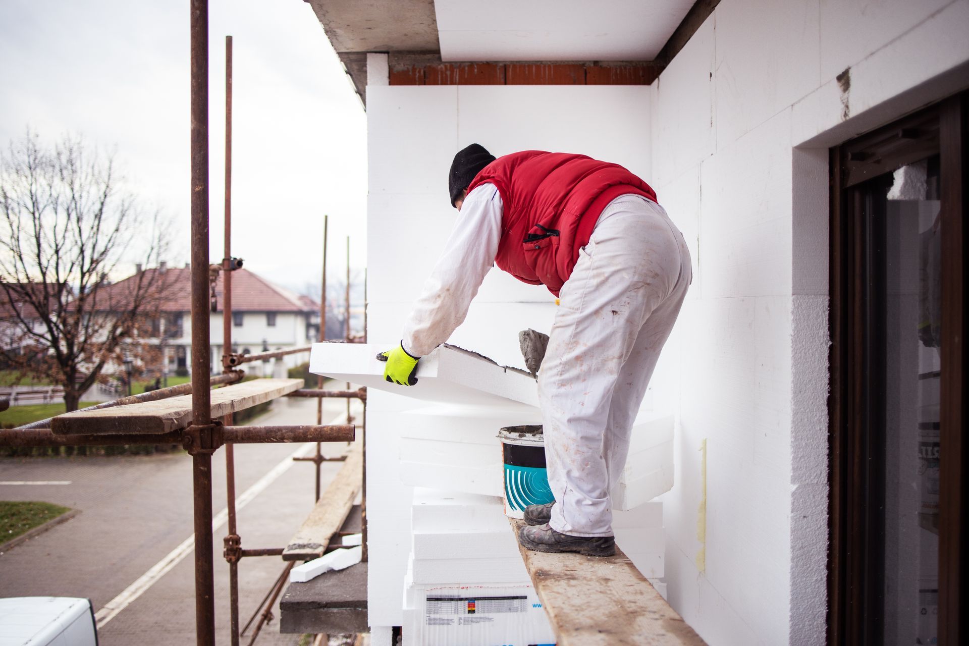 Ouvrier du bâtiment installant de l'isolant sur la façade d'un bâtiment ; mousse blanche, gilet rouge, échafaudage.