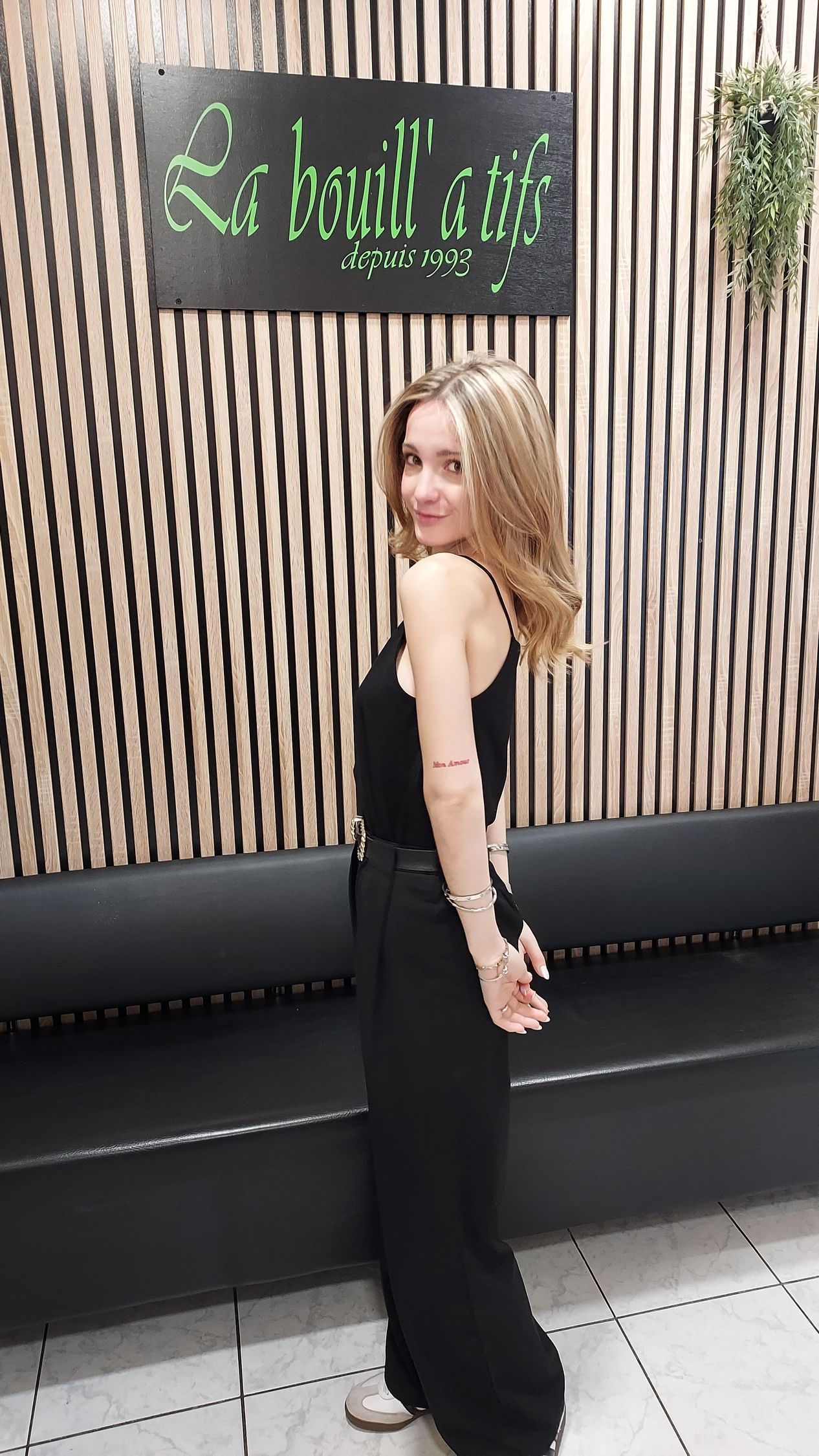 Femme en robe noire, debout de profil devant un mur à lattes de bois et un banc noir.