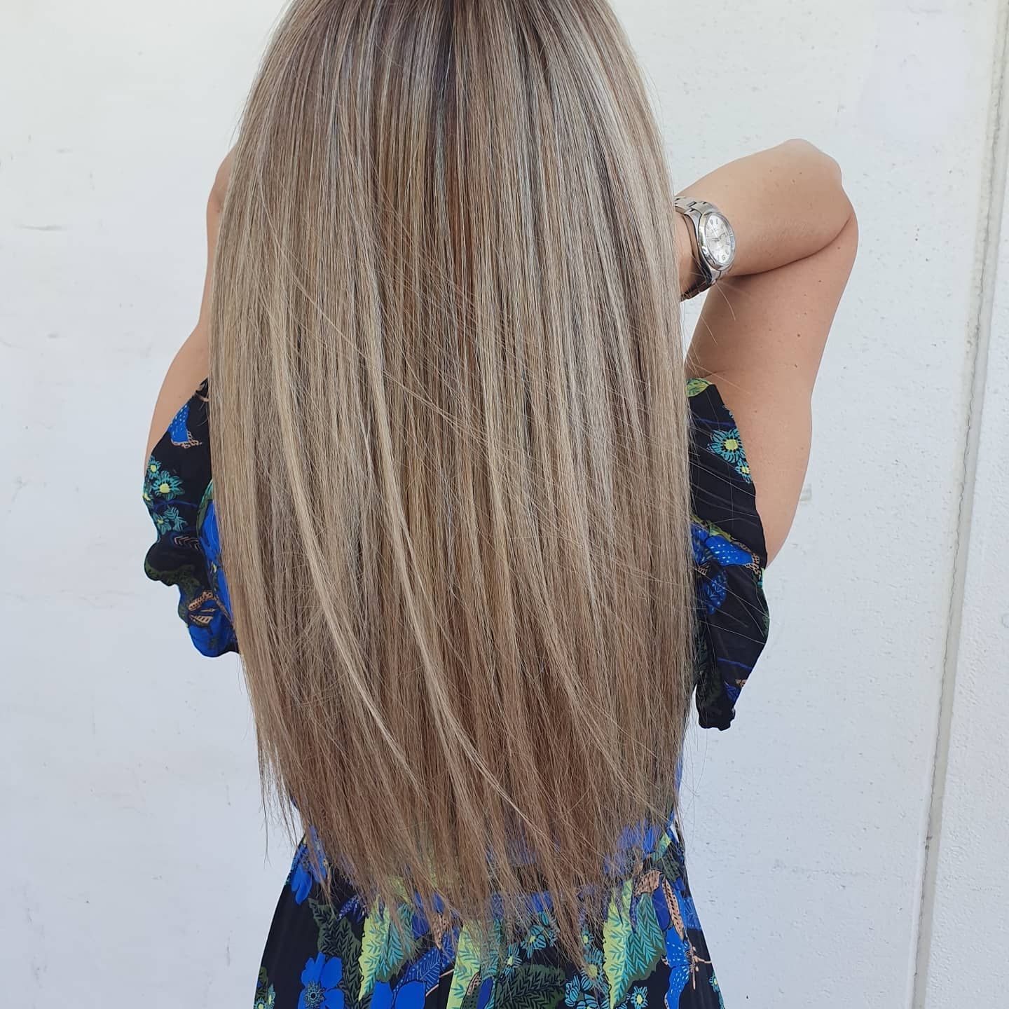 Personne aux longs cheveux blonds, de dos, portant un haut bleu à fleurs, devant un mur blanc