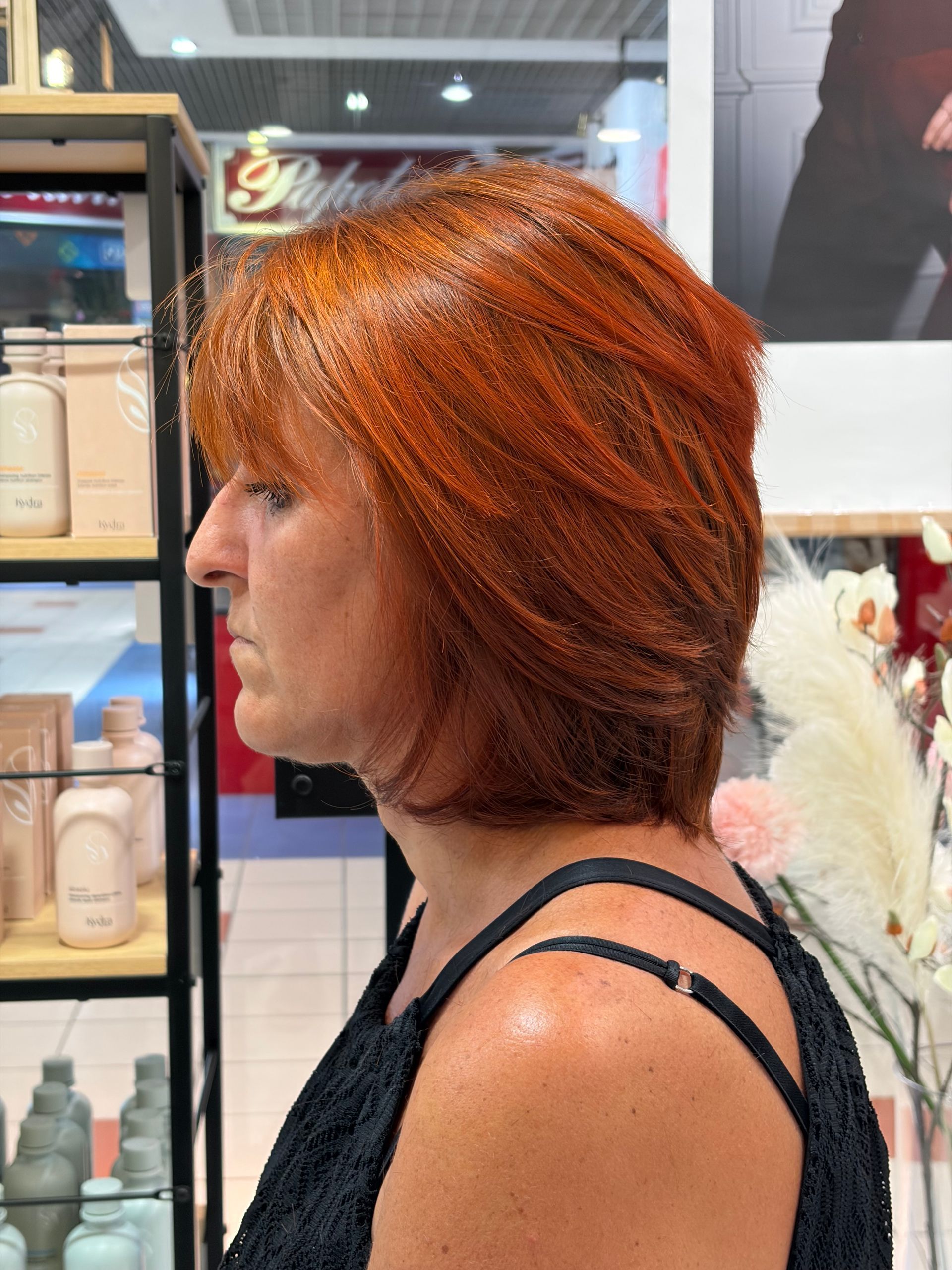 Profil d'une femme aux cheveux courts cuivrés dans un salon de coiffure, portant un haut noir près des étagères de produits