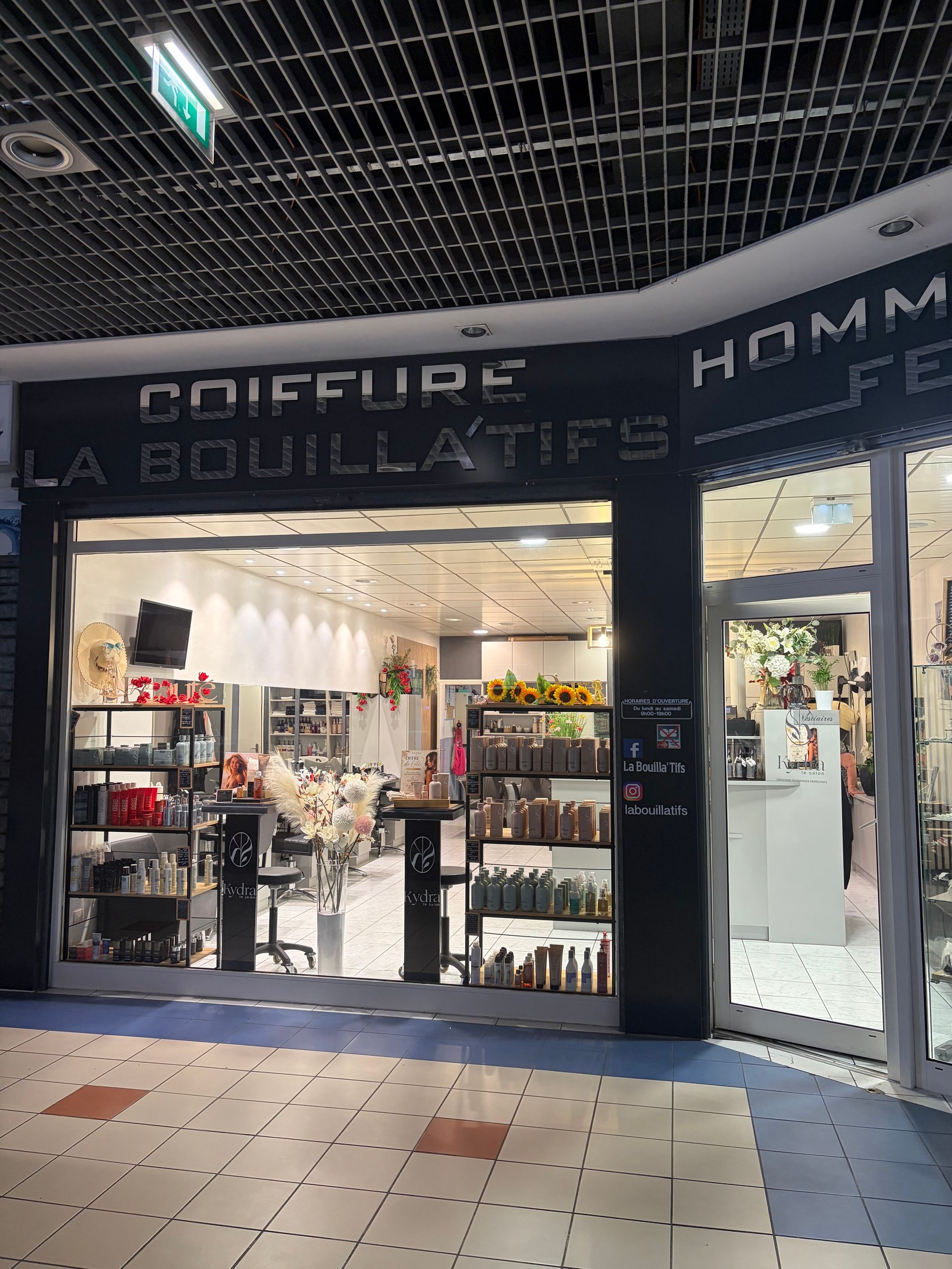 Façade d'un salon de beauté avec vitrines, présentoirs de produits et allée carrelée sous un plafond sombre