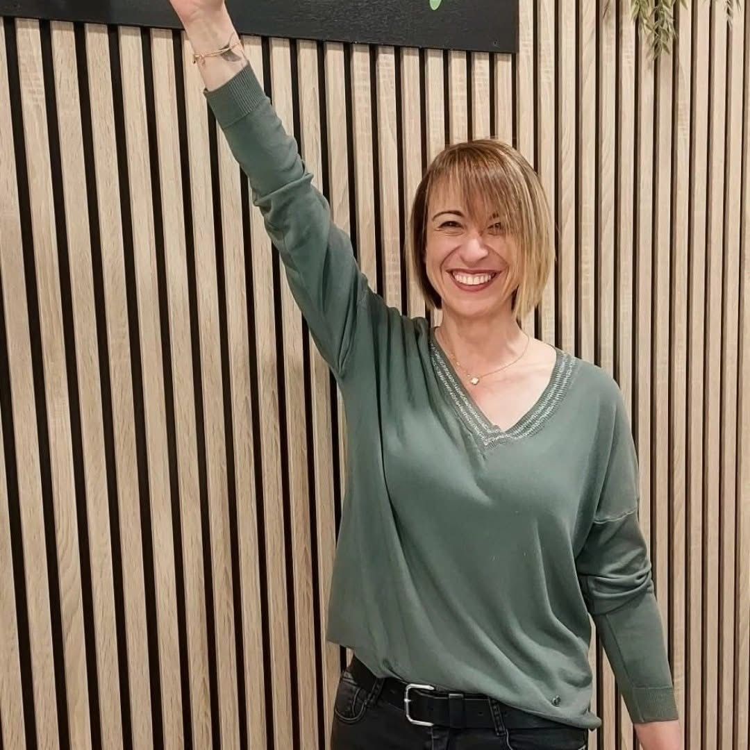 Une personne souriante vêtue d'un haut vert lève un bras devant un mur rayé.