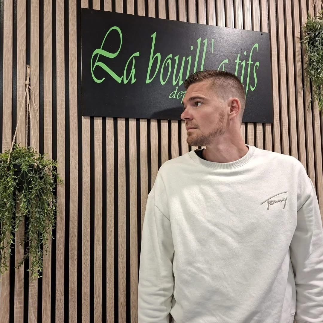 Un homme en sweat-shirt blanc se tient près d'un panneau à inscription verte devant un mur en bois rayé.