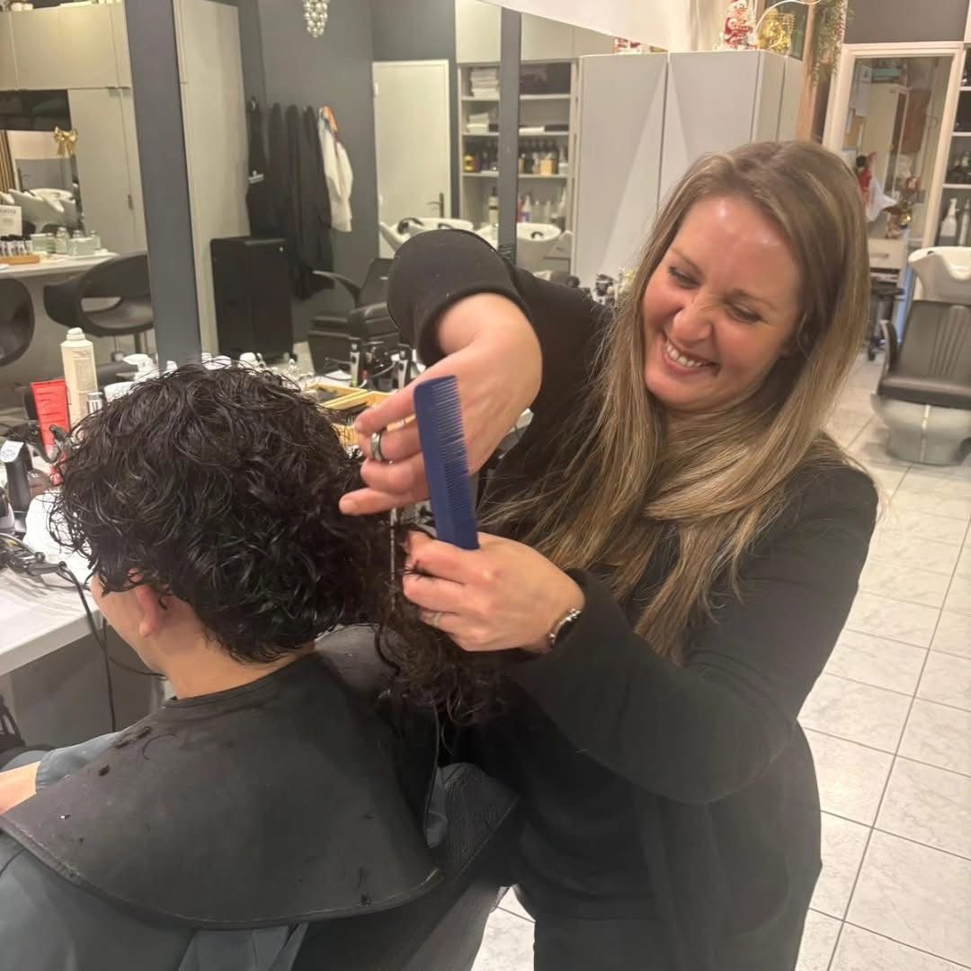 Une coiffeuse coiffe les cheveux bouclés d'une cliente dans un salon, à l'aide d'un peigne et d'un fer à lisser.