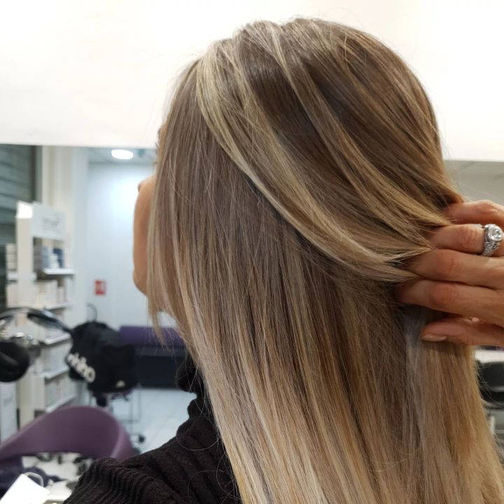 Cheveux blonds coiffés avec de subtils reflets, vus de dos dans un salon de coiffure, une main les ajustant.
