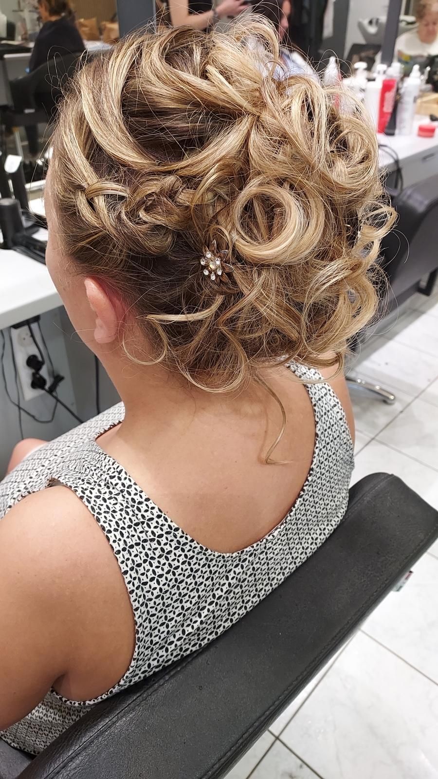 Femme avec une coiffure blonde sophistiquée, assise dans un fauteuil de salon, vue de dos, vêtue d'un haut sans manches à motifs.