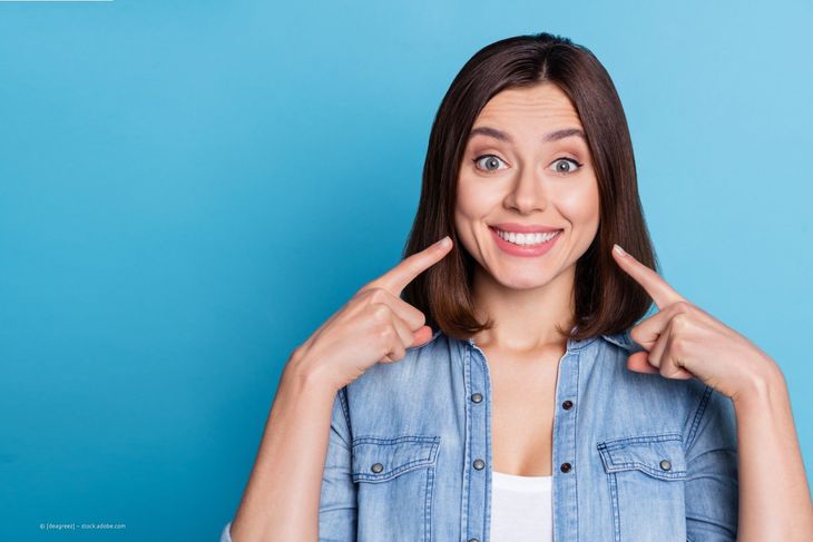Eine lächelnde Frau in einem Jeanshemd deutet vor blauem Hintergrund auf ihre Wangen.
