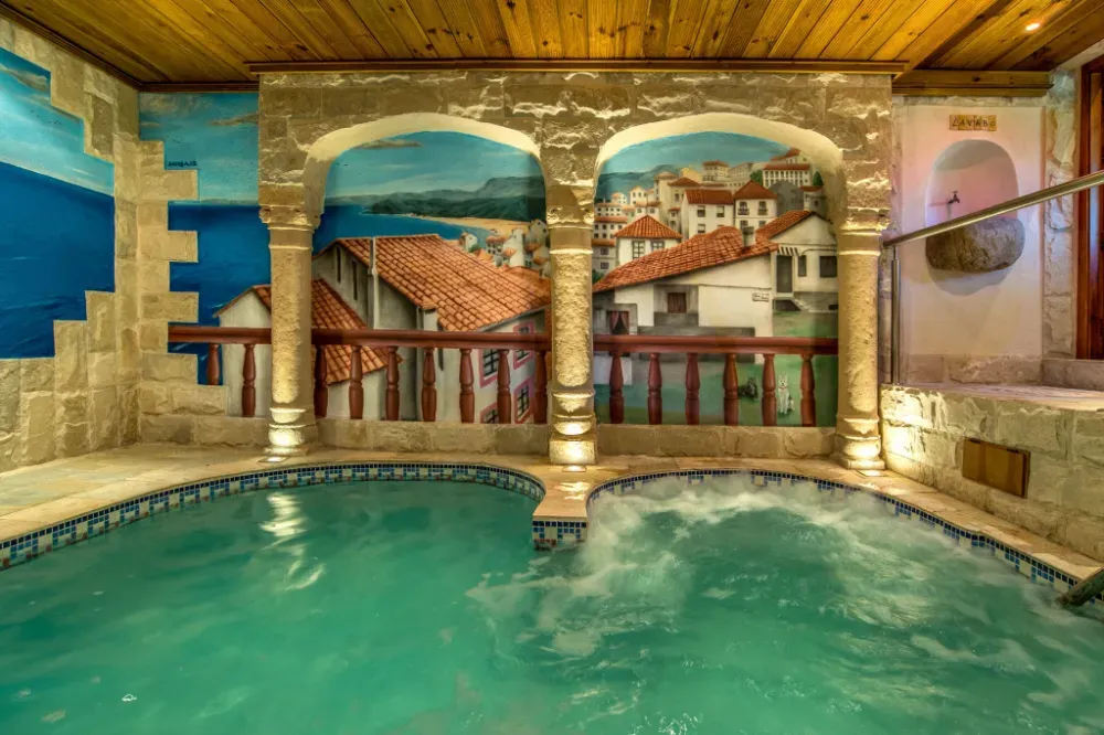 Una bañera de hidromasaje interior con agua turquesa, situada junto a una pared de piedra decorada con un mural de estilo mediterráneo que representa un pueblo costero.