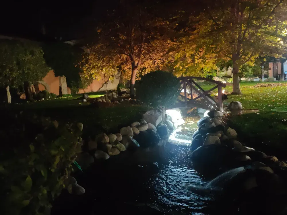 Un pequeño puente de madera con arcos sobre un arroyo iluminado en un jardín por la noche, rodeado de árboles y piedras decorativas.
