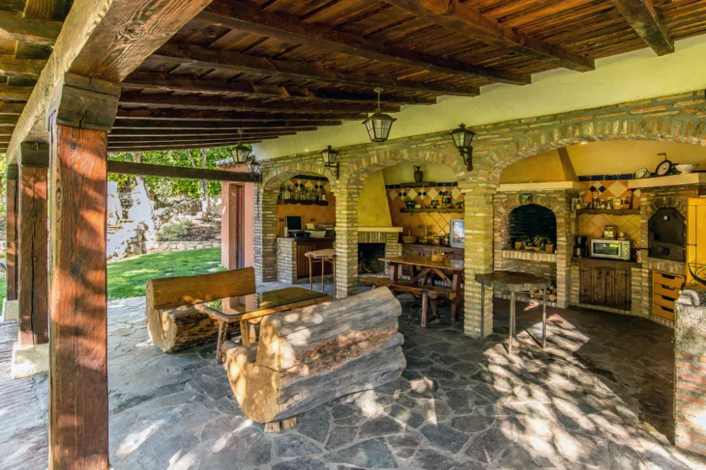 Una cocina y comedor exterior rústicos, con paredes de piedra y techo de madera, que cuenta con bancos de troncos y una parrilla integrada.