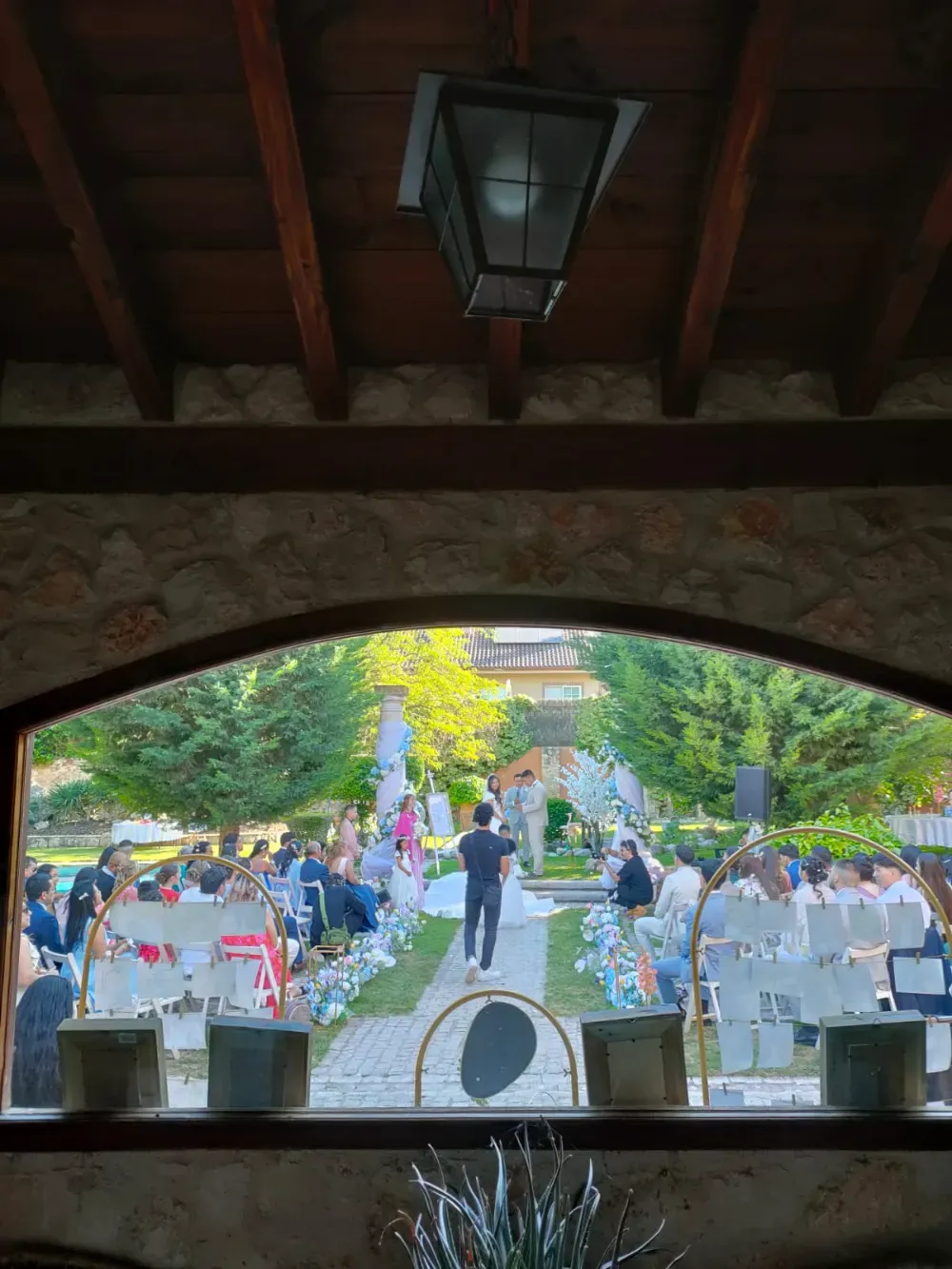 Vista a través de una ventana de piedra arqueada que muestra a los invitados sentados al aire libre en una ceremonia de boda en un día soleado.