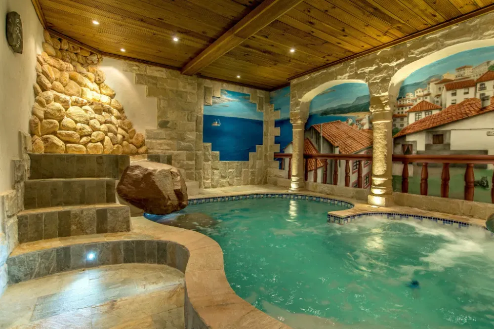 Piscina cubierta con detalles de piedra, elementos rocosos, techo de madera y un mural que representa un pueblo costero.