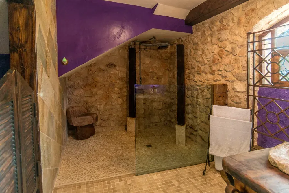 Un baño rústico con paredes de piedra, una ducha a ras de suelo con mampara de cristal, un taburete de madera y un toallero sobre un suelo de baldosas.