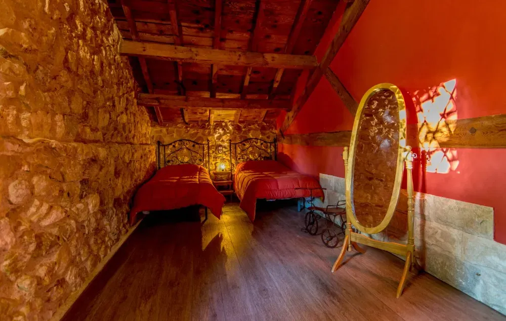 Un dormitorio rústico abuhardillado con dos camas individuales cubiertas con mantas rojas, un espejo ovalado dorado en el suelo y paredes de piedra vista.