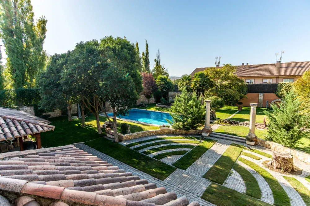 Vista elevada de un jardín paisajístico con piscina azul, columnas de piedra y un diseño geométrico en el césped.