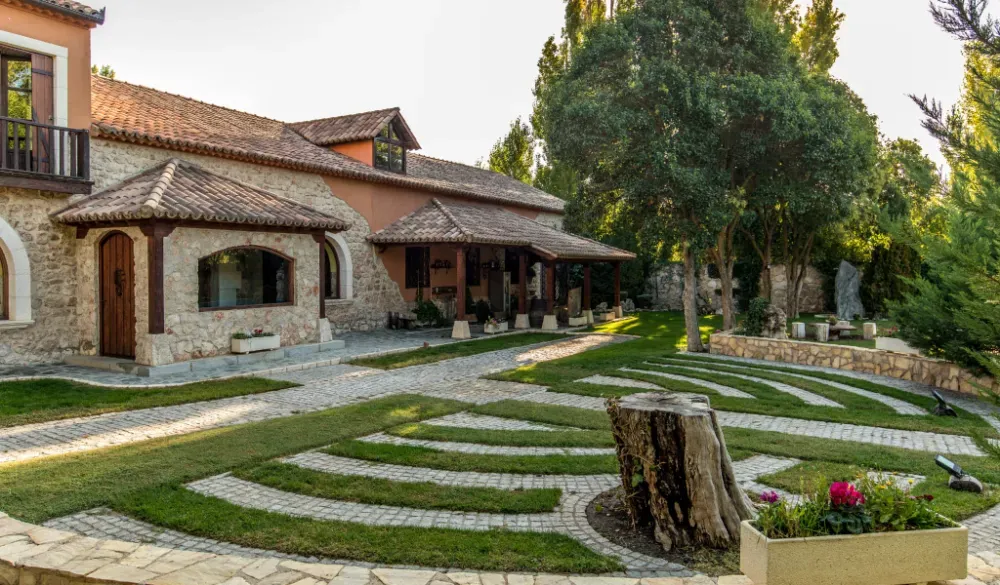 Una casa de piedra con tejado de terracota se alza junto a un jardín curvo y laberíntico, pavimentado con piedra, sobre un césped.