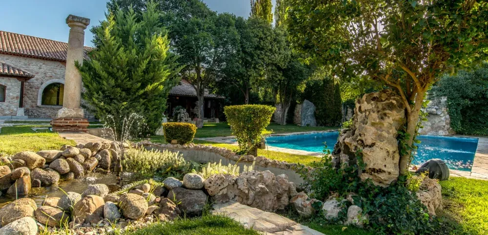 Un jardín con una fuente de piedra, un pilar decorativo, una piscina y frondosos árboles cerca de un edificio de piedra.