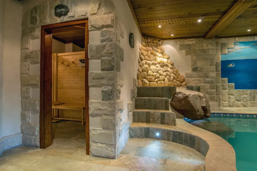 Una sala de spa con sauna de madera, escaleras de piedra y una piscina cubierta de color turquesa con una gran roca decorativa.