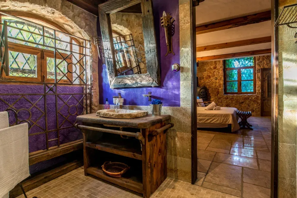 Un baño con paredes de piedra, una pared de color morado, un lavabo de madera y un espejo, con vistas a un dormitorio con cama y suelo de piedra.