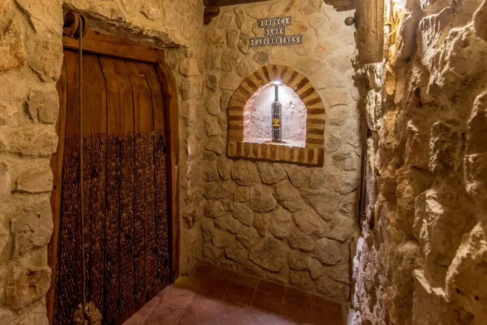 Un pasillo rústico de piedra con una puerta de madera a la izquierda y una alcoba arqueada que exhibe una botella de vino a la derecha.