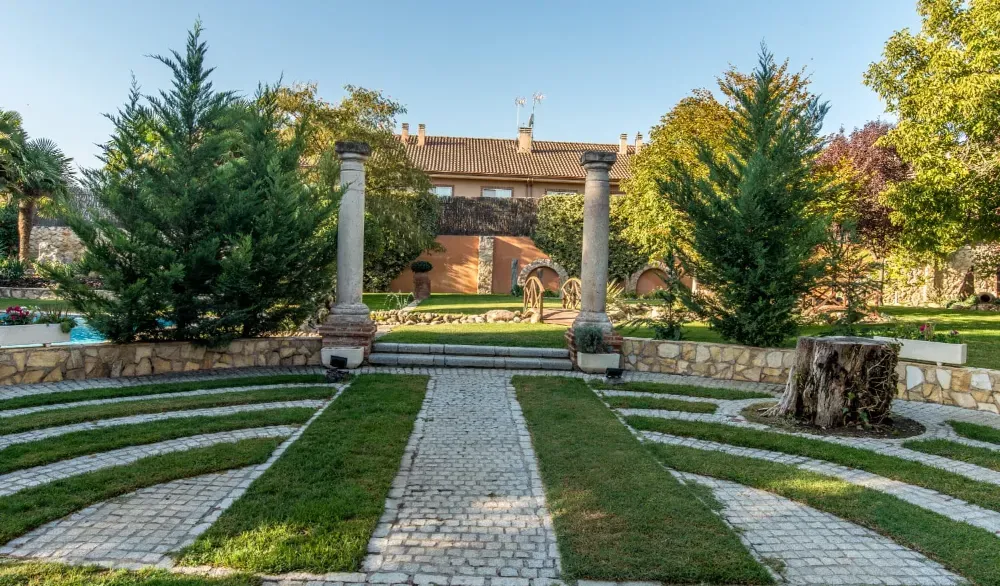 Un sendero empedrado conduce a dos altas columnas en un jardín paisajístico con césped bien cuidado y árboles de hoja perenne.