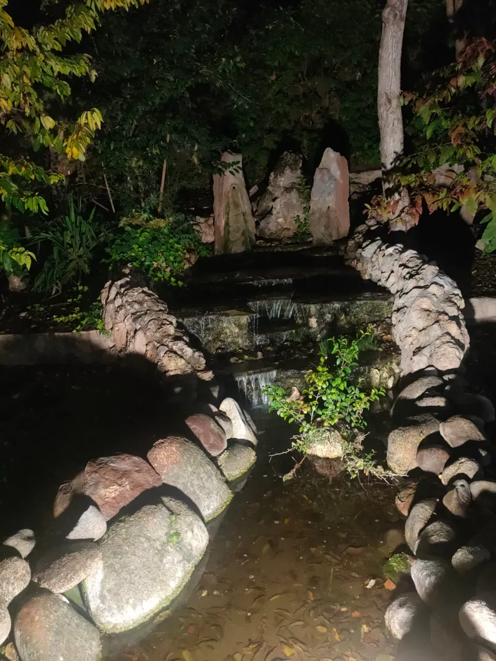 Una pequeña cascada de piedra en un jardín por la noche, iluminada por una luz tenue, que desemboca en un estanque bordeado de rocas.