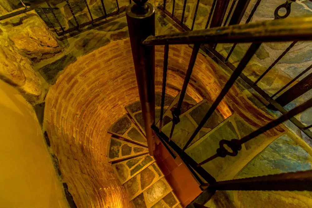 Vista desde arriba de una escalera de piedra en espiral con barandilla de metal oscuro, rodeada de muros curvos de ladrillo texturizado.