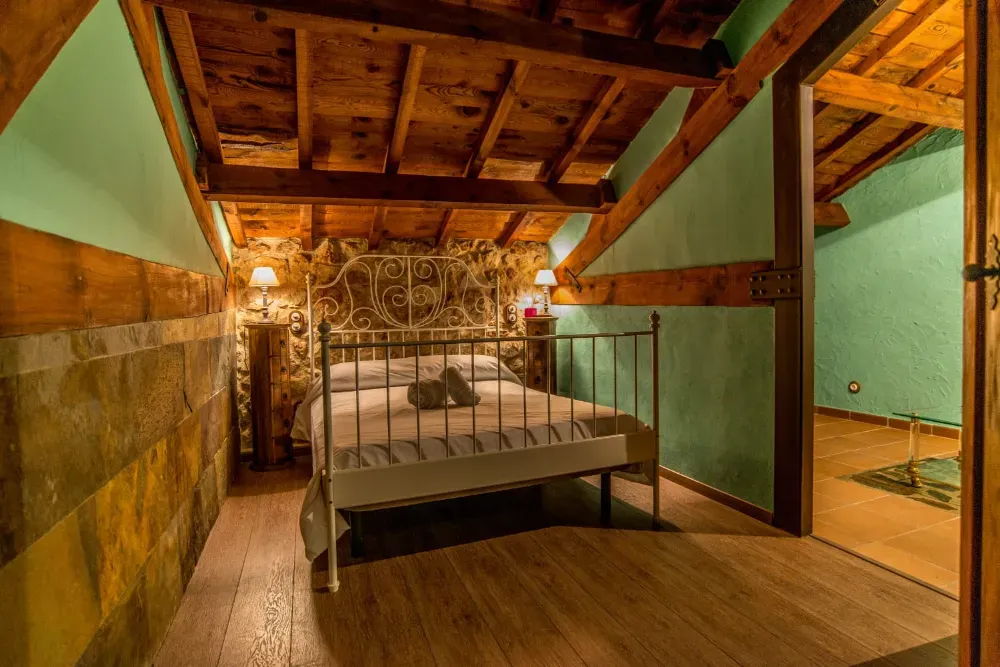 Un dormitorio rústico abuhardillado cuenta con una estructura de cama de metal, paredes de color verde claro, un techo con vigas de madera y suelo de madera.