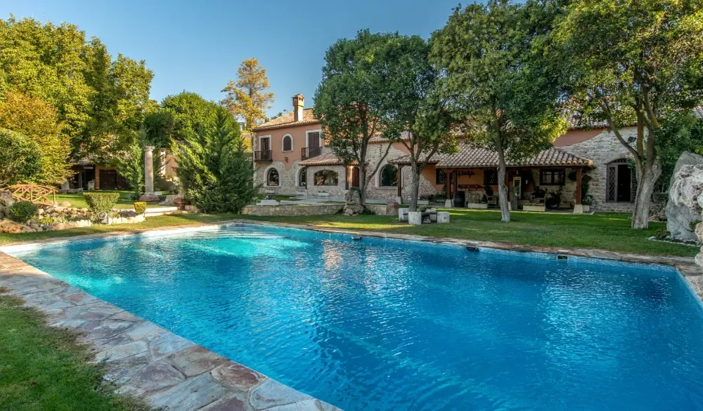 Una piscina de color azul brillante con bordes de piedra se ubica frente a una extensa villa de estilo mediterráneo de varias plantas.