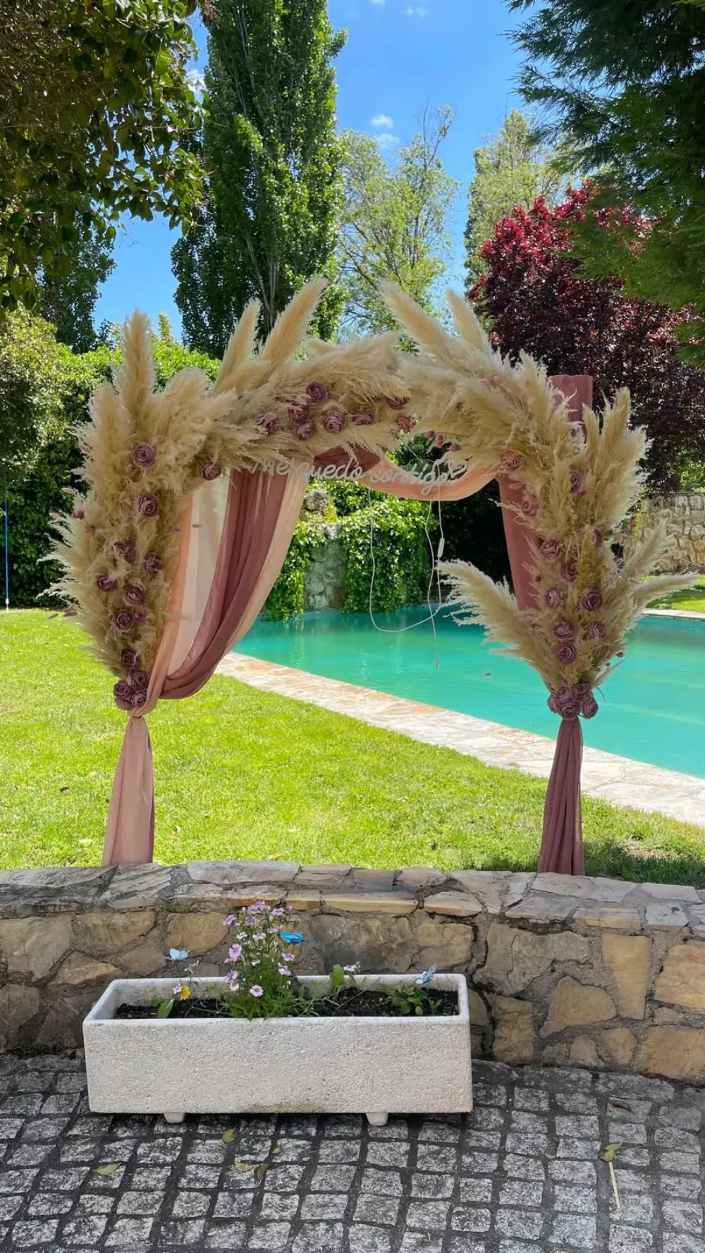 Un arco floral al aire libre, decorado con hierba de la pampa y tela rosa empolvada, se alza frente a una piscina y un césped.
